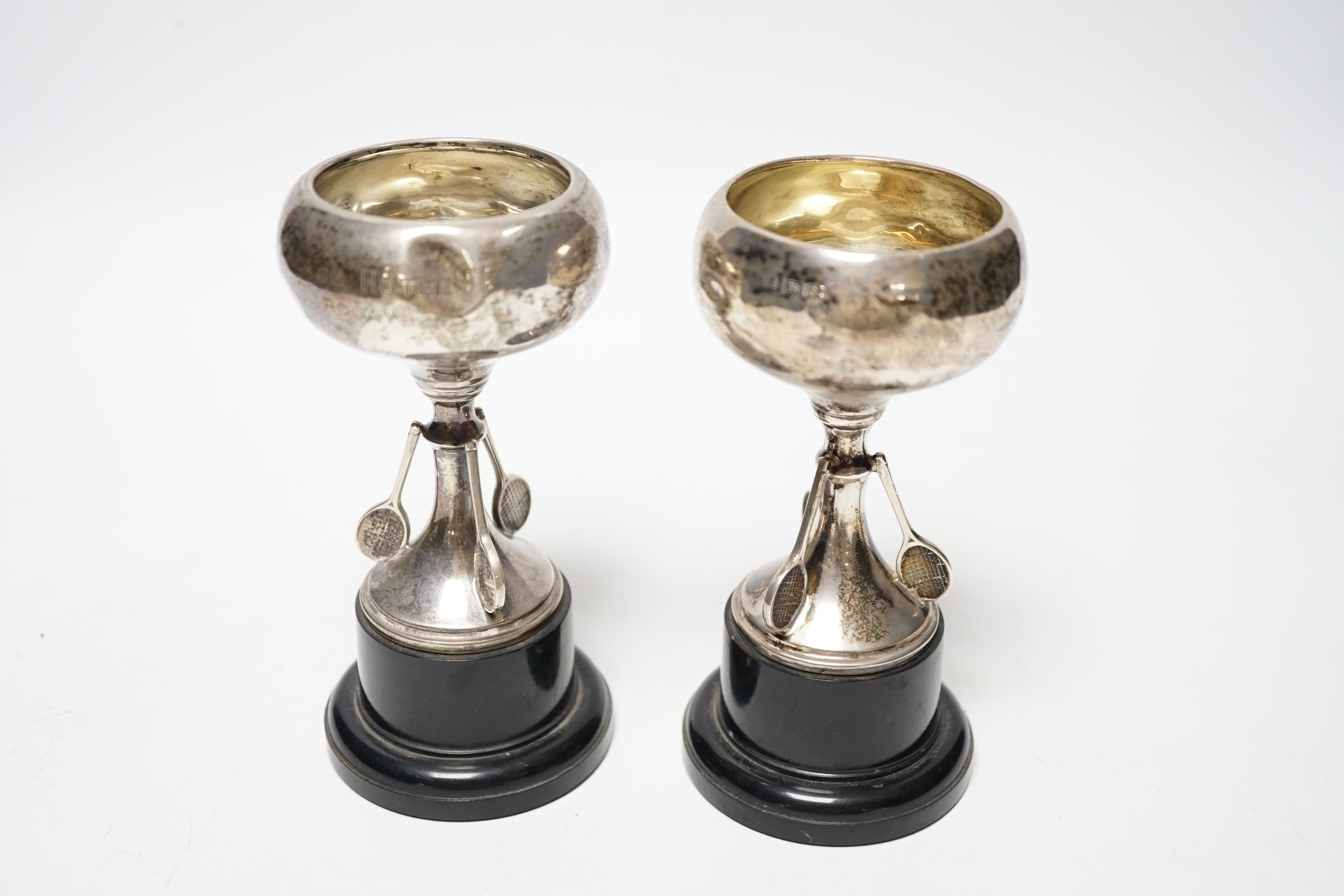 A set of three small George V silver tennis related trophy cups, with racket supports and engraved inscriptions, E & J Leek, 92mm, two with plinth bases.